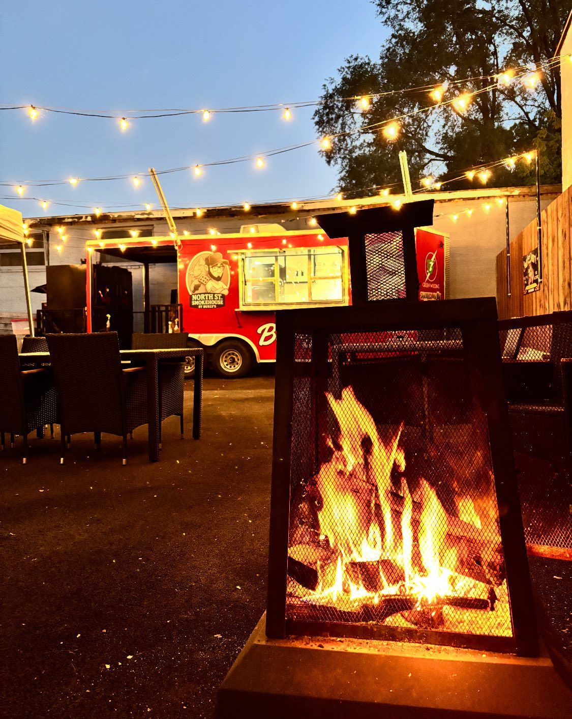 Outdoor seating area with lit fire pit and food truck in the background, strung with lights.