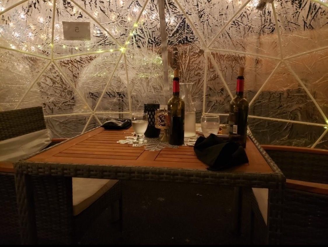 Inside dining igloo with table, chairs, wine bottles, and string lights.