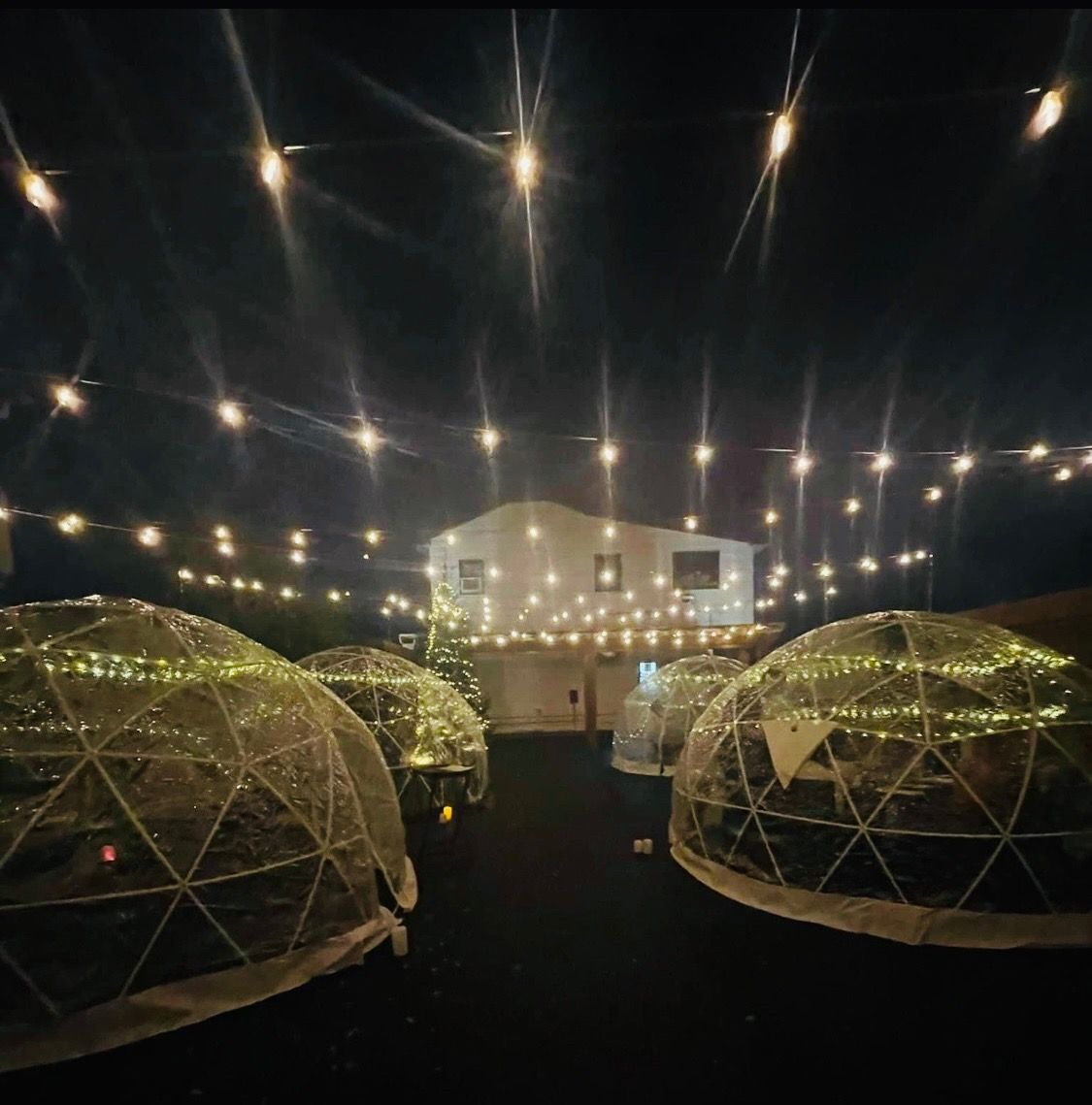 Outdoor dining area with lit igloo-shaped domes under string lights, a white building in the background.