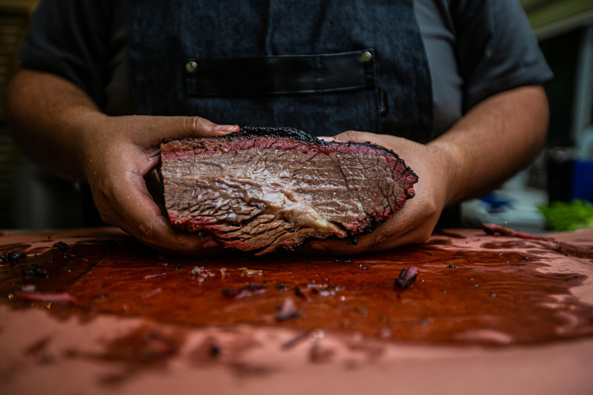 Person holding a large, sliced piece of smoked brisket on a butcher's table.