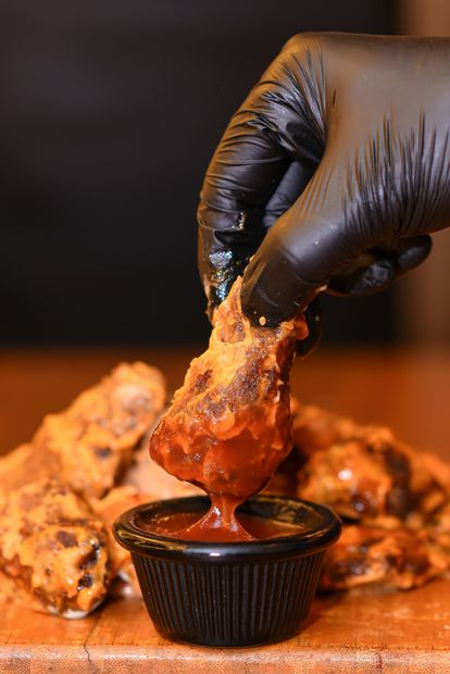 A hand in a black glove dipping a fried chicken wing into a small container of red sauce.