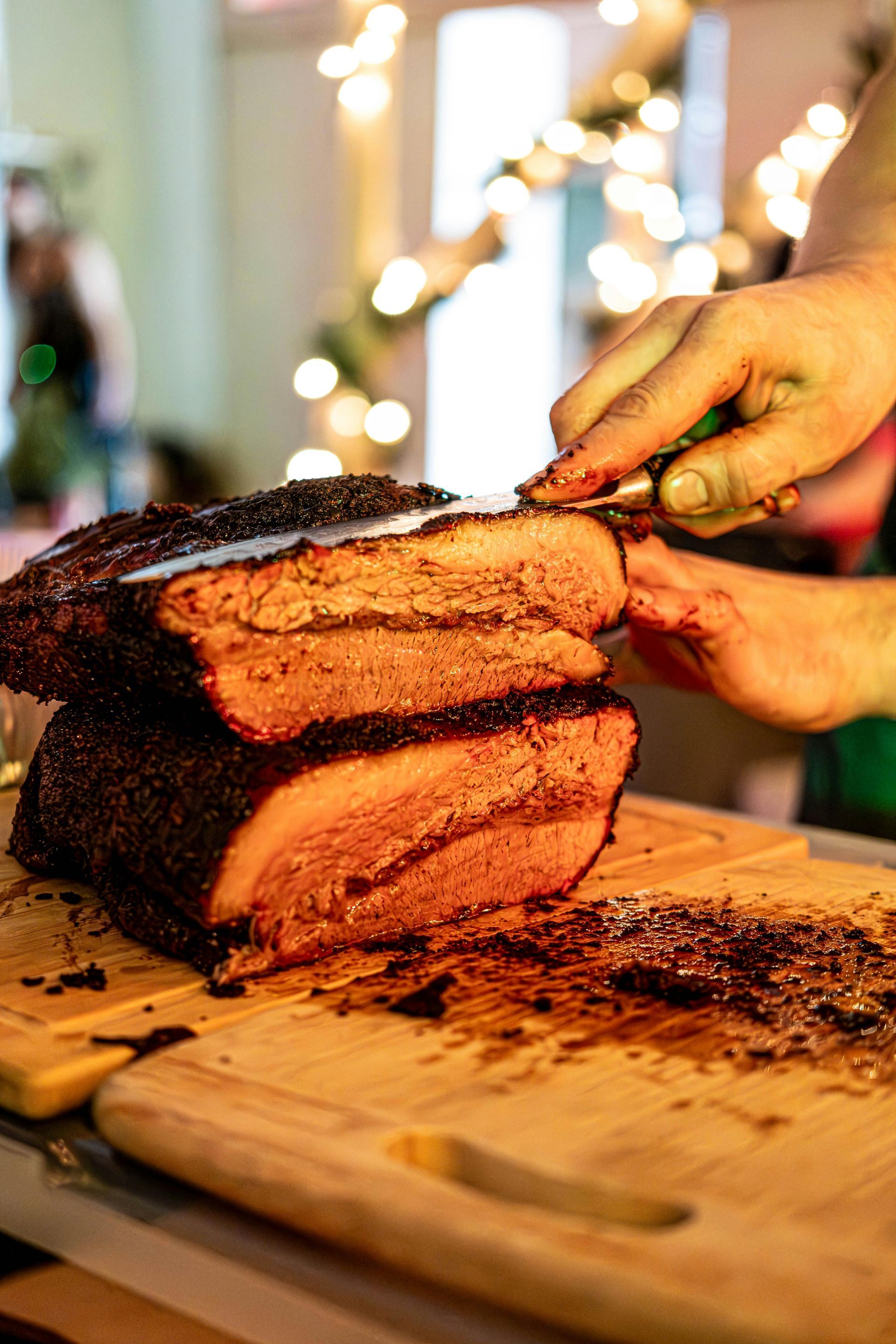 Person slicing smoked brisket on a wooden cutting board; indoor setting with festive lights.