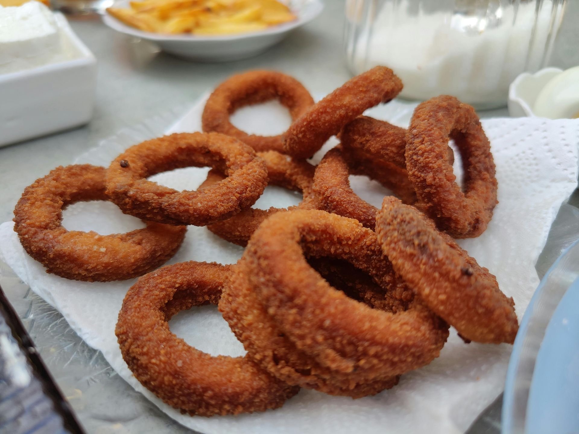 Pile of golden-brown onion rings on a white napkin, with sides of food in the background.