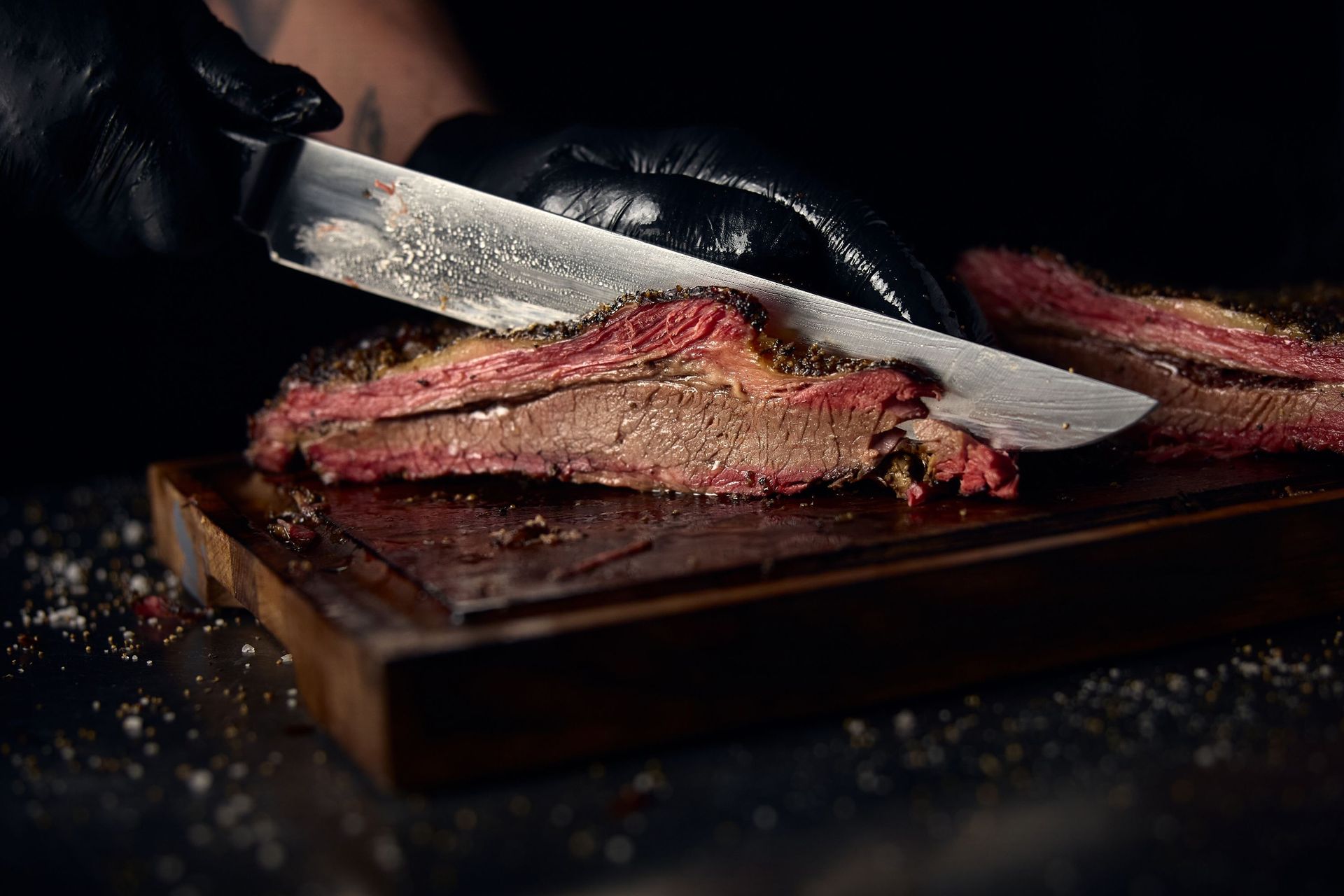 Person wearing black gloves slicing brisket on a wooden cutting board with a knife.