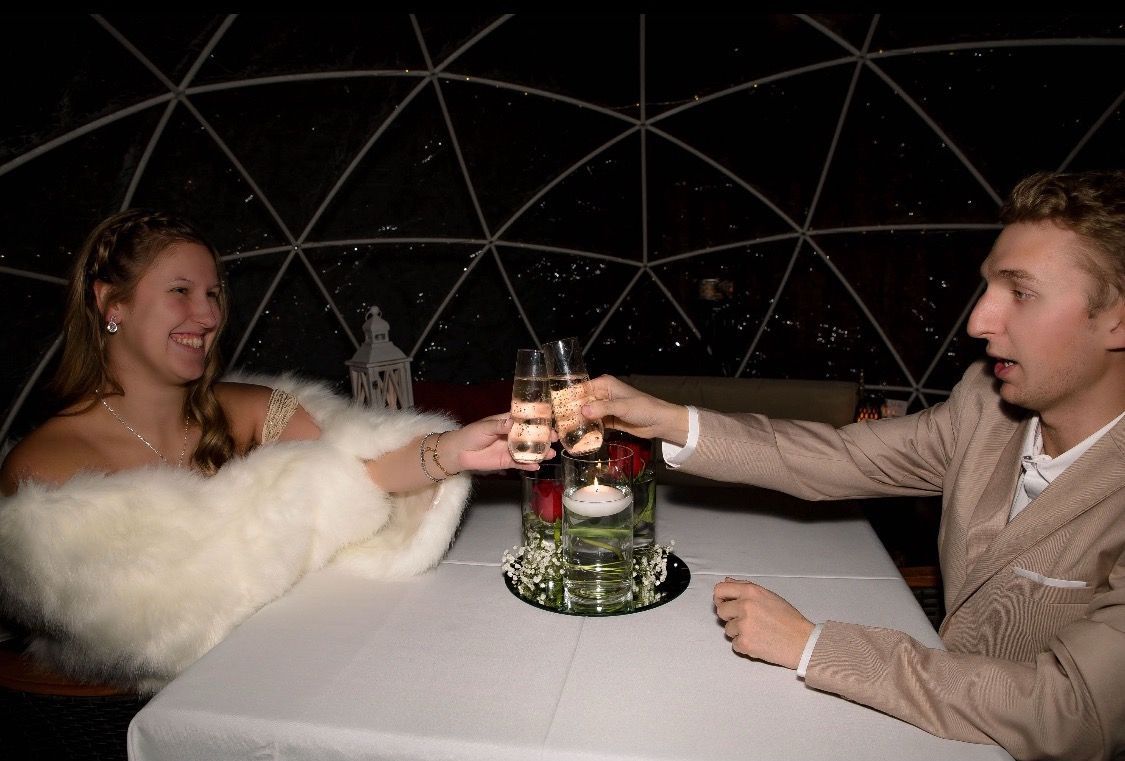 Couple toasting champagne at a table inside a geodesic dome, nighttime setting.