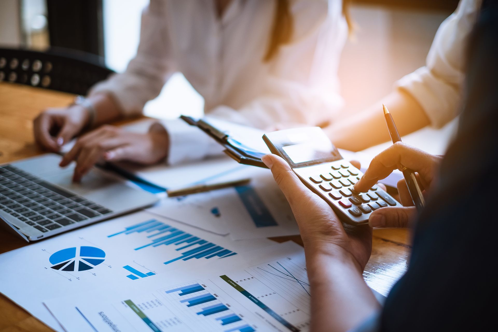 Hands using a calculator over financial charts and a laptop at a table