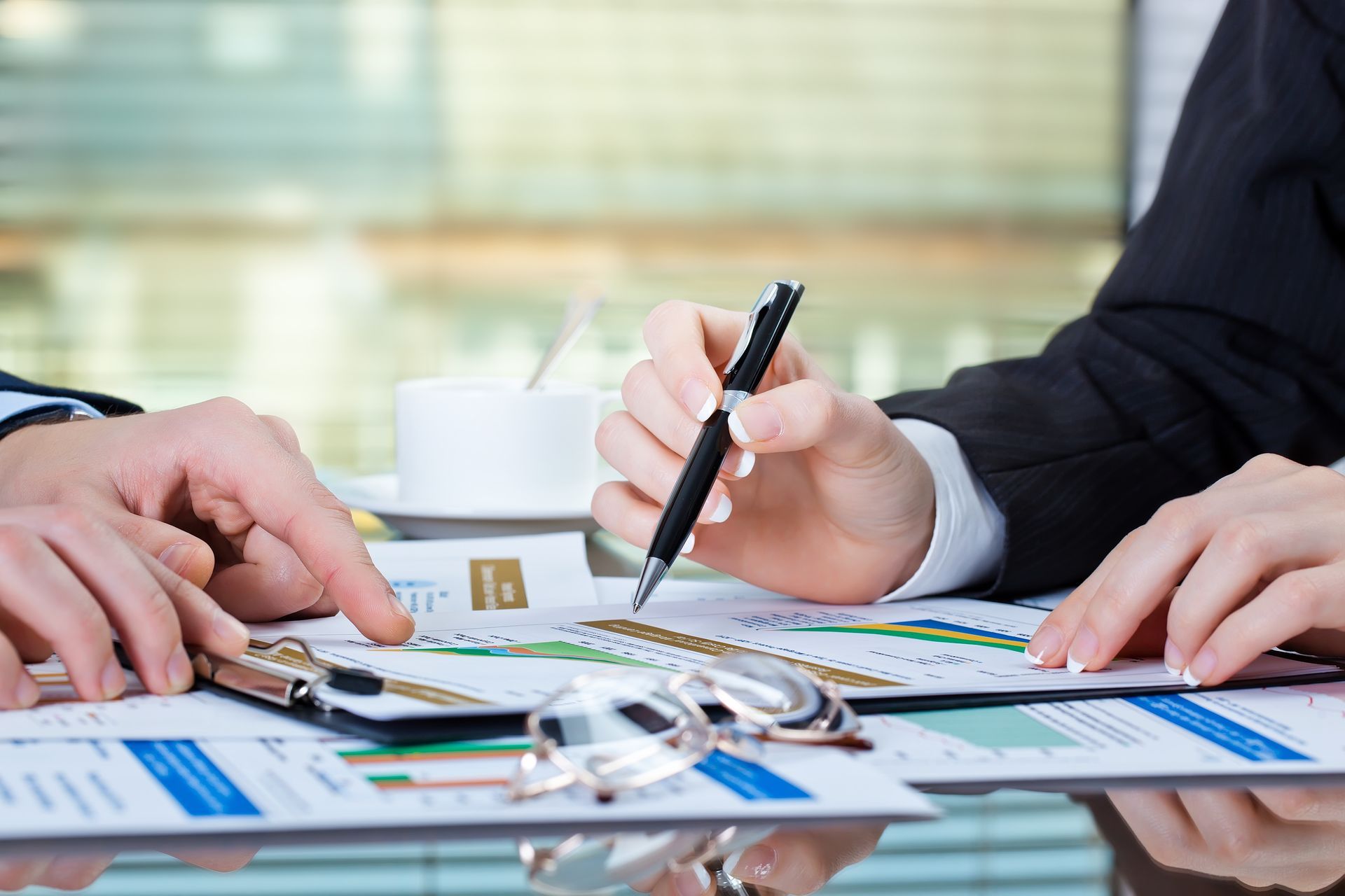 Hands pointing to financial charts during a meeting. Pen, glasses, and coffee cup on the table.