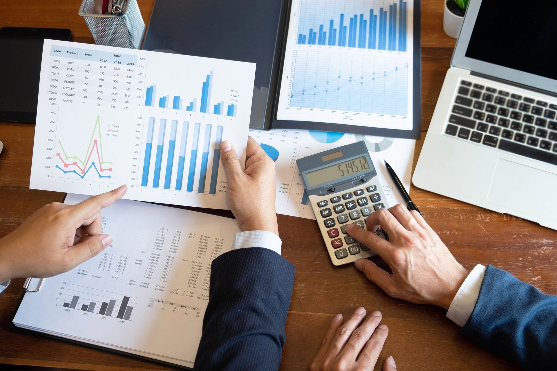 Two people analyzing financial data, using a calculator and laptop, on a wooden desk.