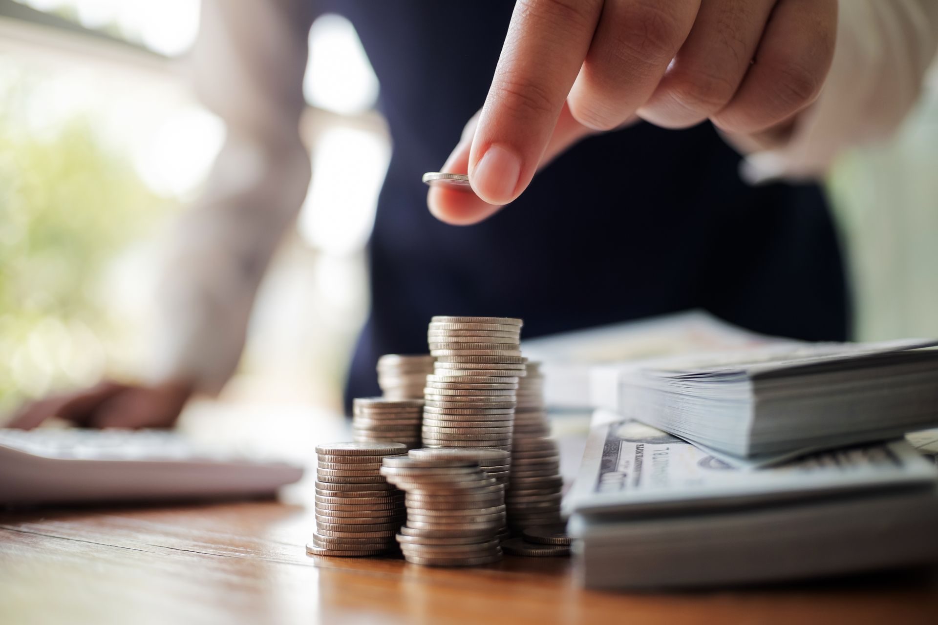 Person adding coin to a stack of coins on a table with money and calculator