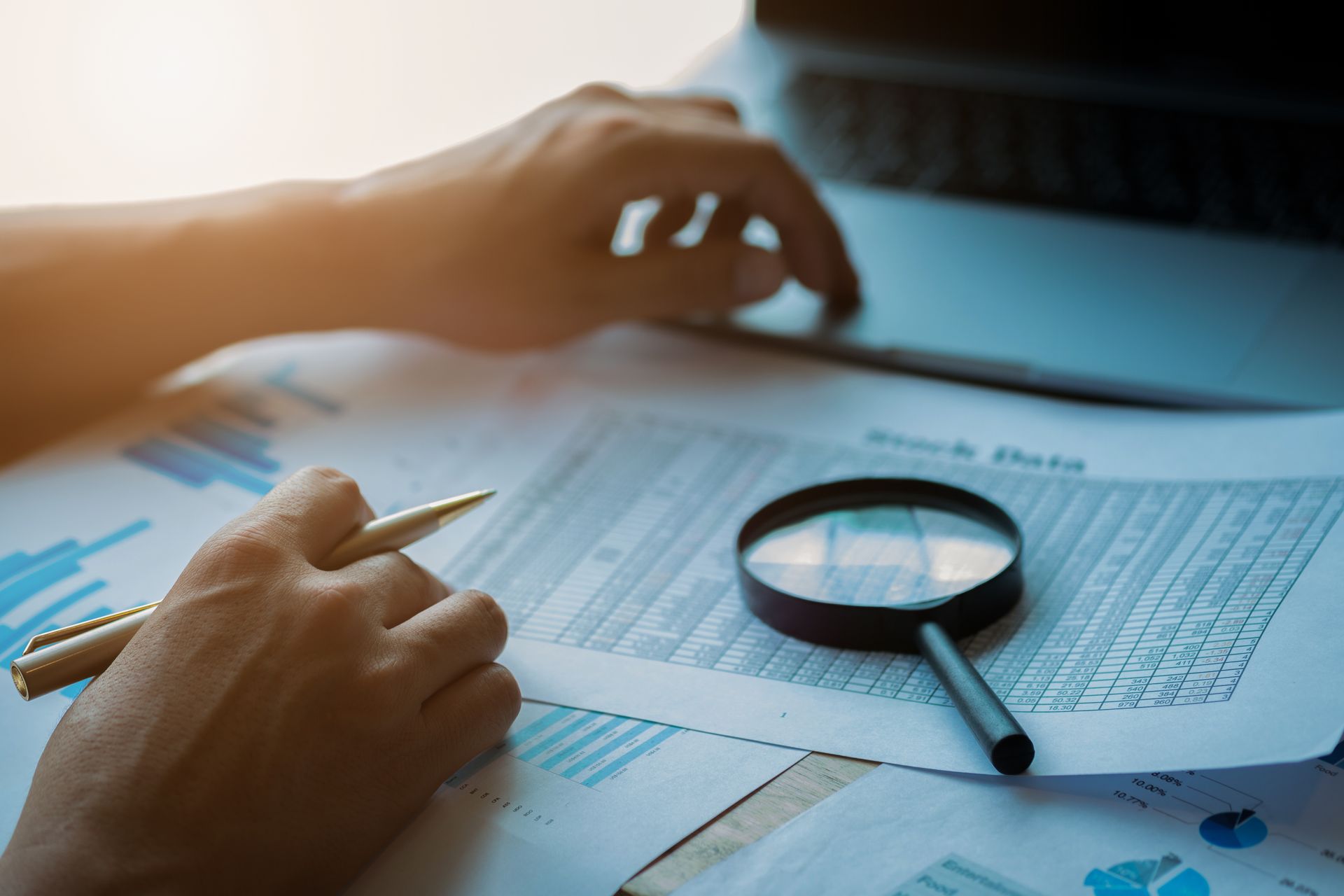 Person examining financial documents with a magnifying glass and laptop