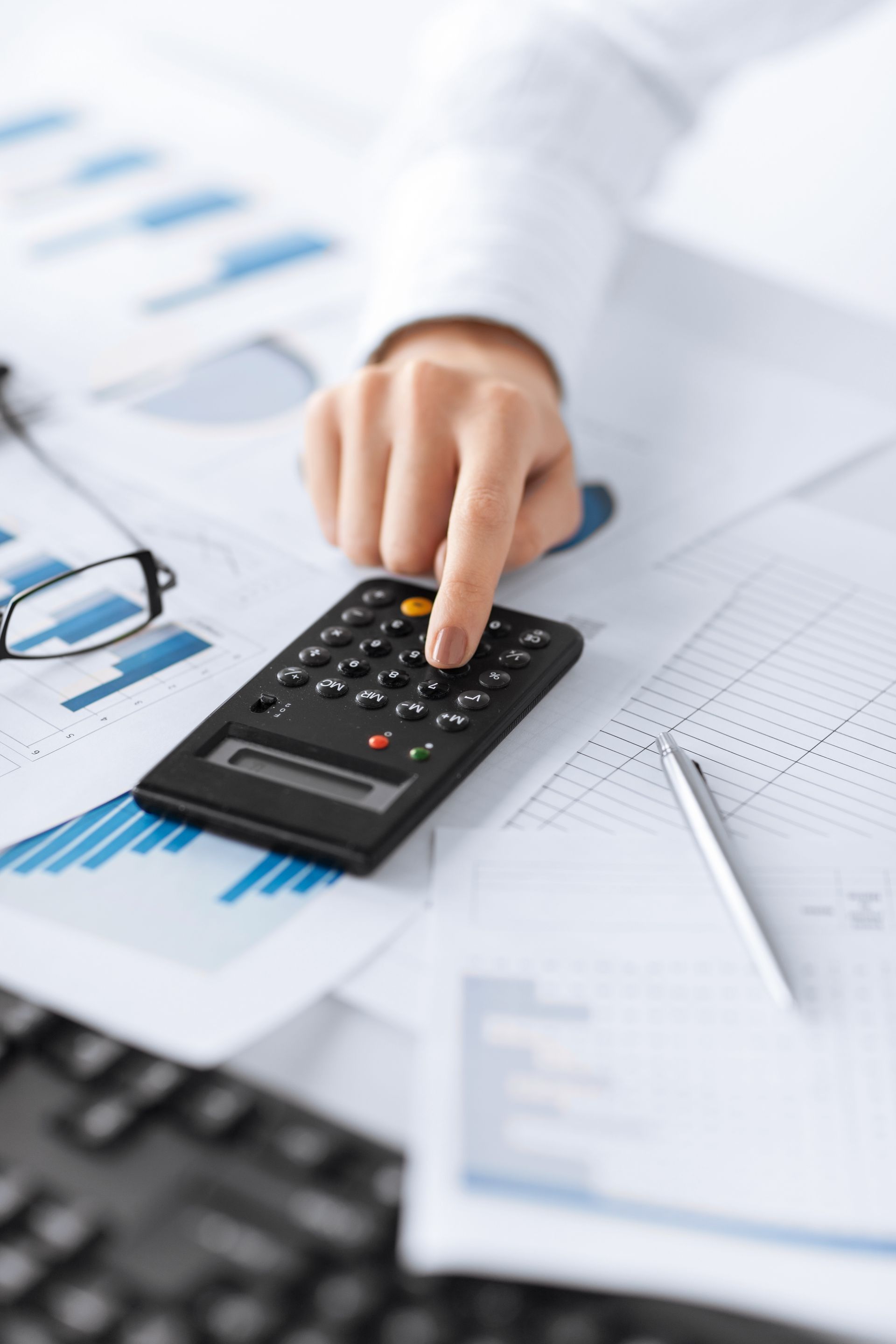 Person's hand using a calculator on a desk with charts, papers, glasses, and a pen