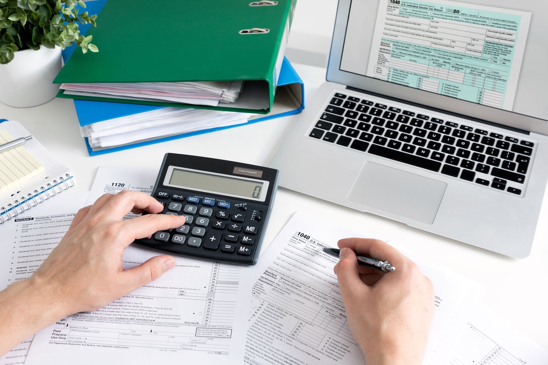 Hands using a calculator and pen next to documents and a laptop displaying a tax form