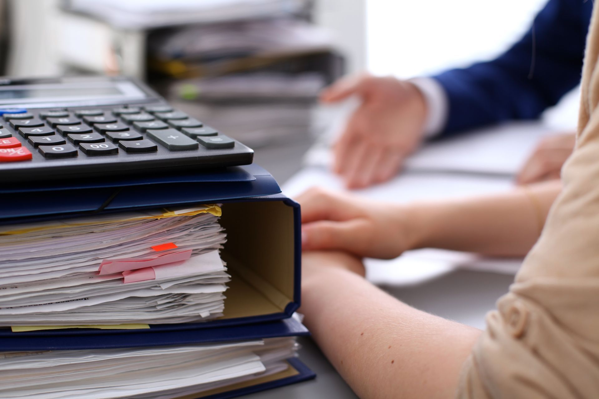Stack of paperwork with a calculator and people discussing documents at a desk