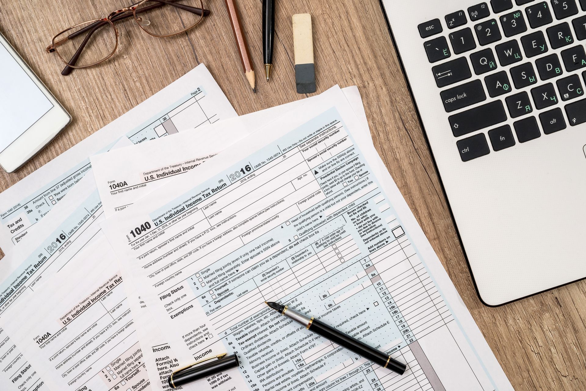 Tax forms, laptop, pens, eraser, glasses, and phone on a wooden desk