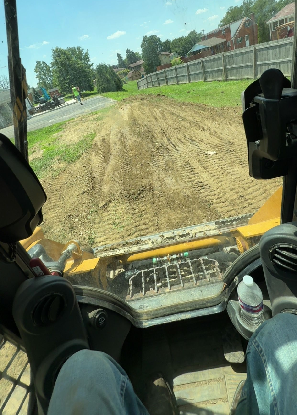 View from a skid steer, looking over a graded dirt area toward a street and buildings.