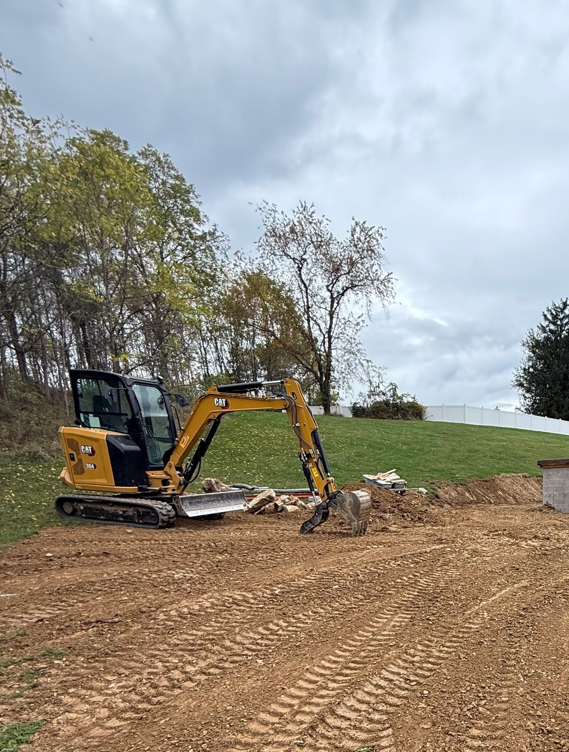 Yellow excavator digging in a dirt area, with a grassy hill and cloudy sky in the background.