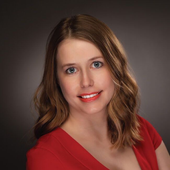Woman with light brown hair in a red top smiles at the camera against a gray background.