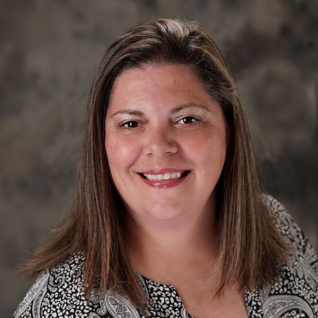 Woman with brown hair smiling, wearing a black and white paisley print top, in front of a gray backdrop.