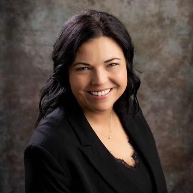 Woman in black blazer smiling at the camera, dark hair against a textured background.