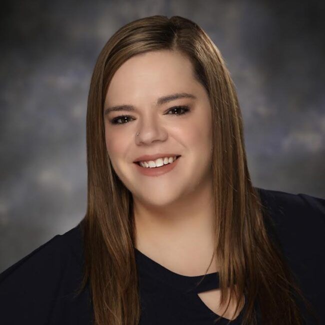Woman with long, brown hair smiles at the camera, wearing a dark blue top against a cloudy background.