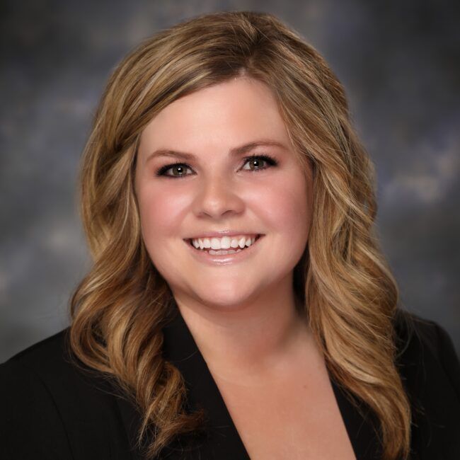Woman with blonde hair smiling, wearing a black blazer, against a blurred background.