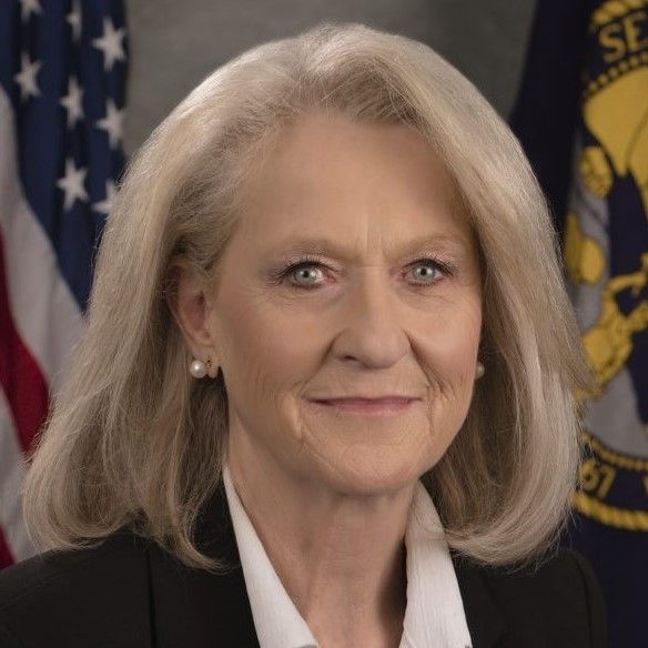 Woman with fair skin and light hair, wearing a black blazer. American flag in background.