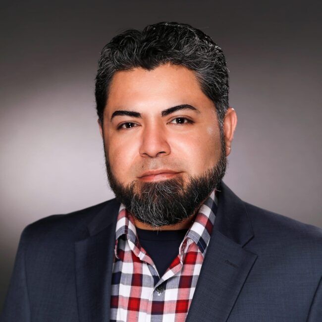 Man in a blazer and plaid shirt, with a trimmed beard, looking directly at the viewer.