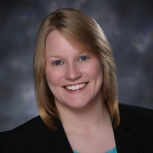 Woman with blonde hair smiling, wearing a black blazer and light blue top against a blurred background.