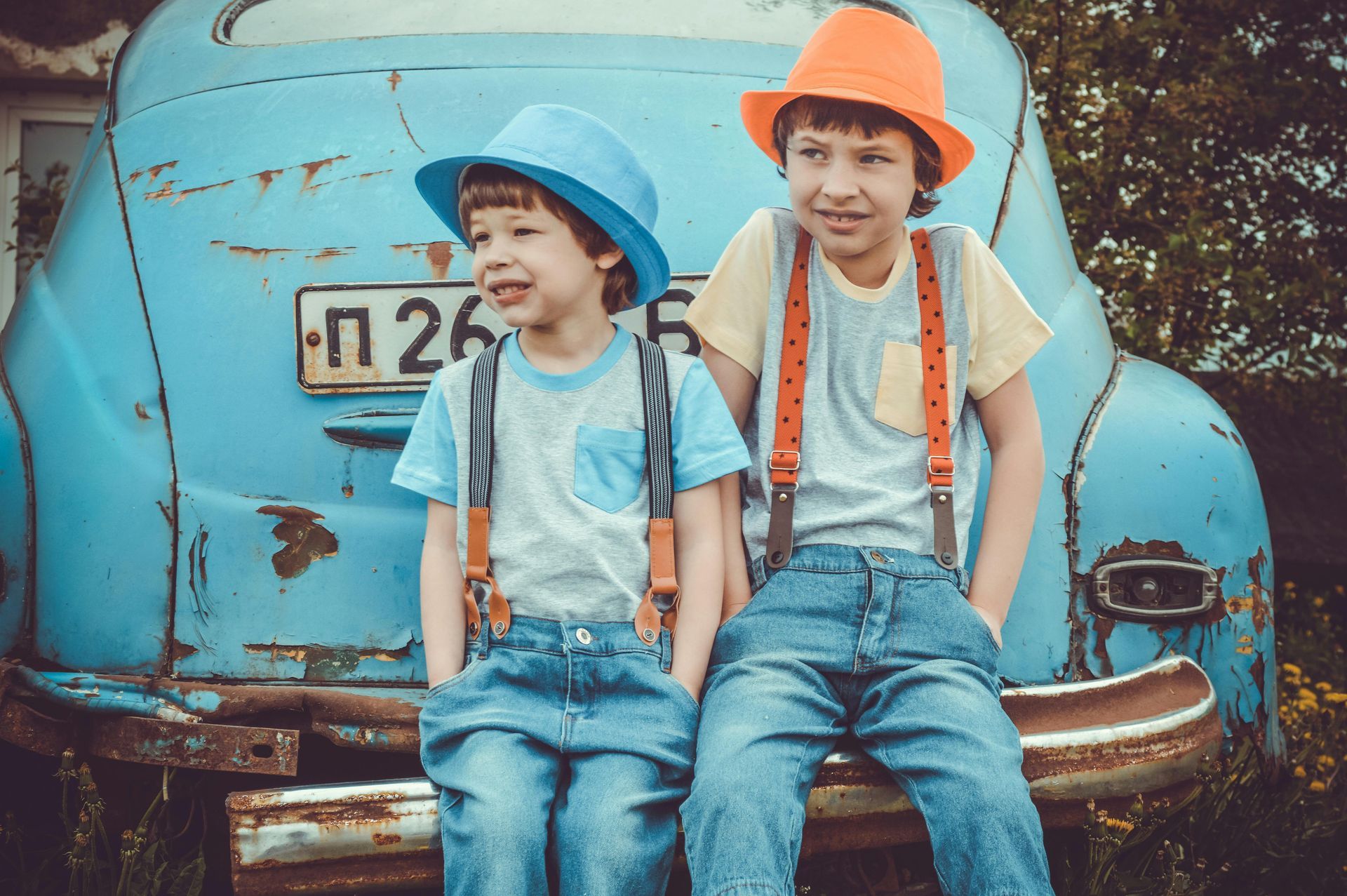 Two children in hats and suspenders pose in front of a blue vintage car.