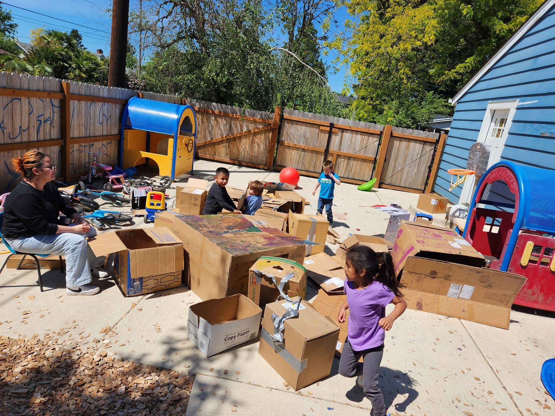 A little girl in a purple shirt is standing in front of a pile of cardboard boxes.