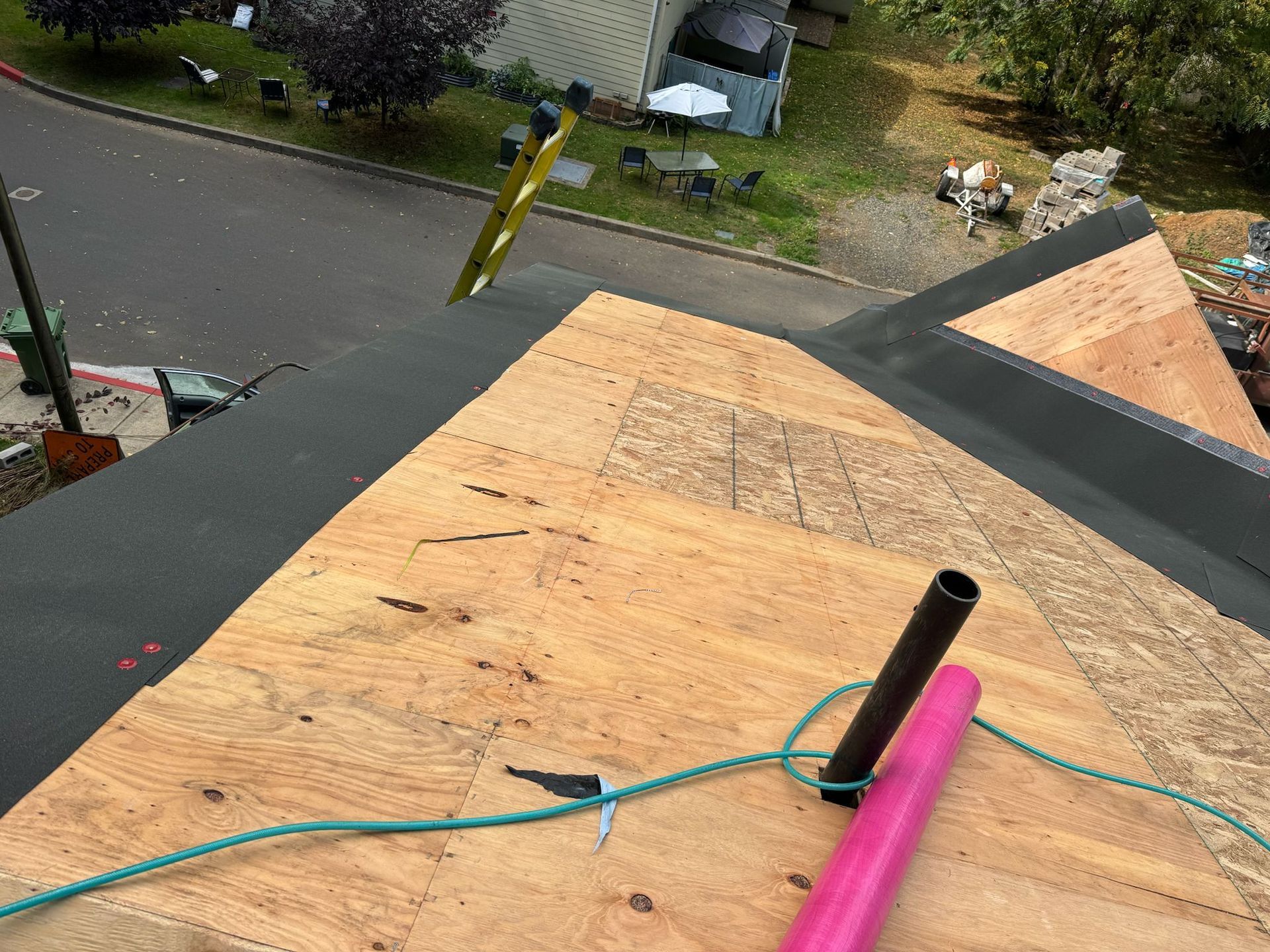 Rooftop view of a partially shingled roof with a black pipe, pink tube, and a green rope.