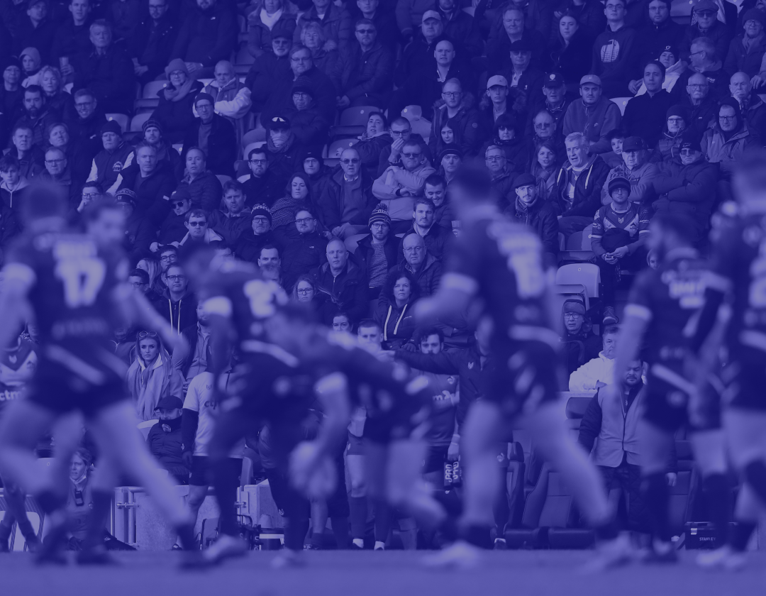 Rugby game in stadium with blurred players in foreground and crowd in blue tint.