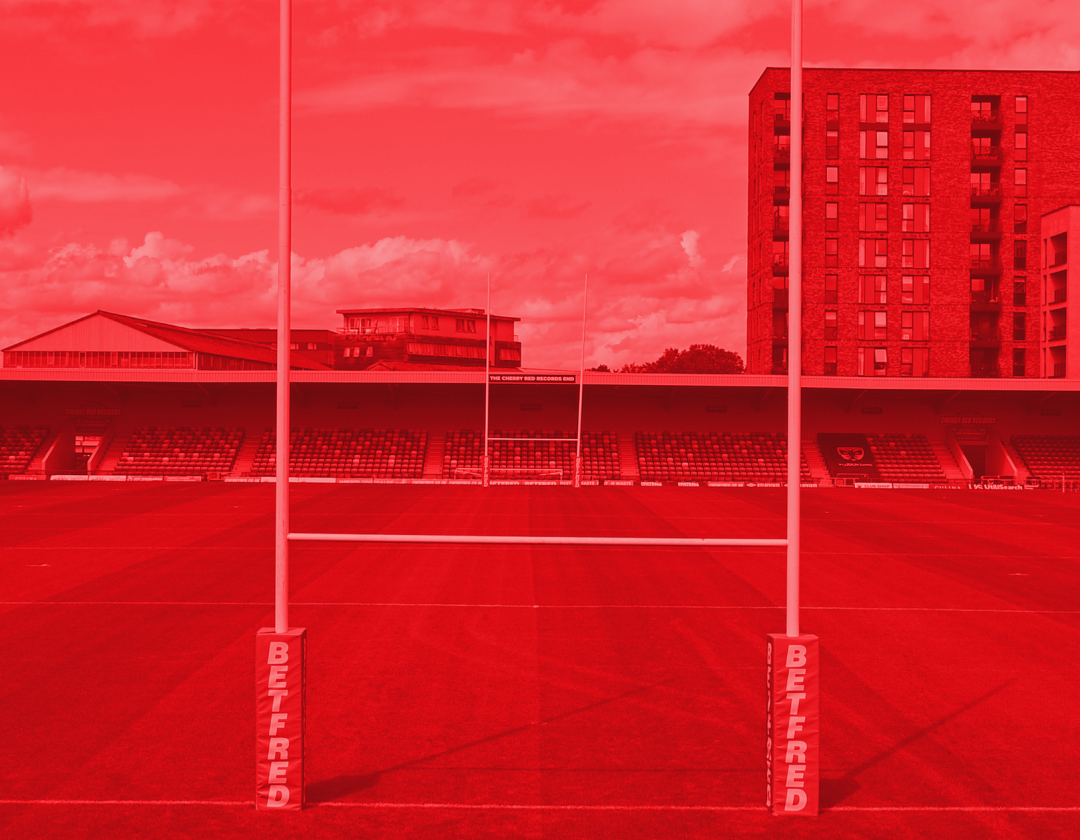 Red-tinted view of a rugby field with goalposts, bleachers, and buildings in the background.