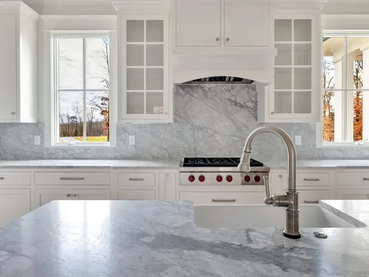 Bright white kitchen with marble island, farmhouse sink, stainless stove, and large windows.