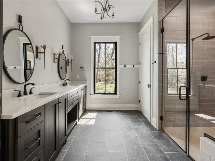 Modern bathroom with dark vanity, large mirrors, gray tile floor, window, and glass shower enclosure