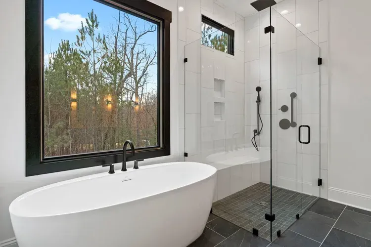 Modern bathroom with freestanding tub, black fixtures, glass shower, and large window overlooking trees