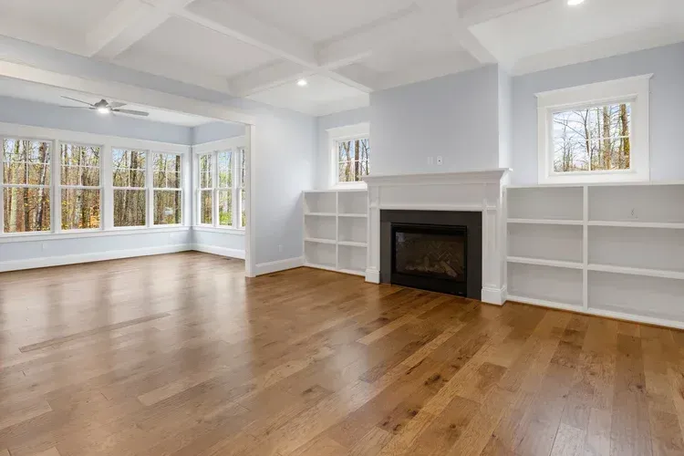 Bright empty living room with hardwood floors, white walls, large windows, and a fireplace