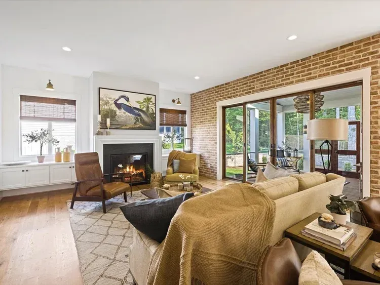 Bright living room with fireplace, beige sofas, wood floor, large windows, and brick accent wall