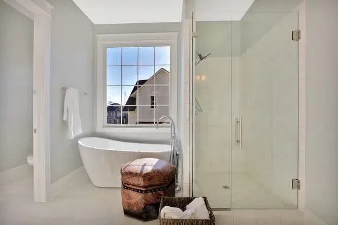 Bright modern bathroom with freestanding tub, glass shower, and window view of a house