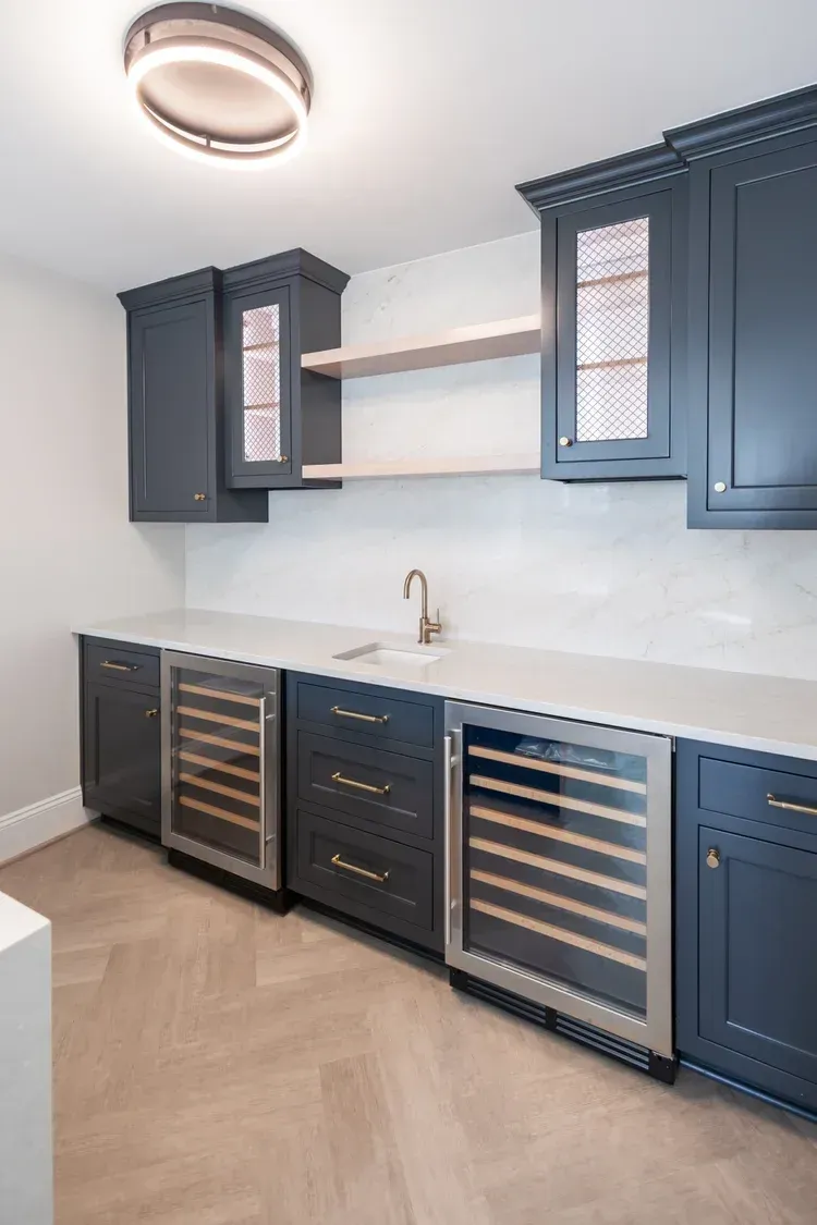 Built-in blue-gray cabinets and wine fridges in a modern wet bar with white backsplash and counters