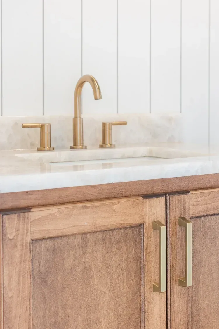 Gold faucet and fixtures over a white bathroom sink with wood vanity and vertical wall paneling