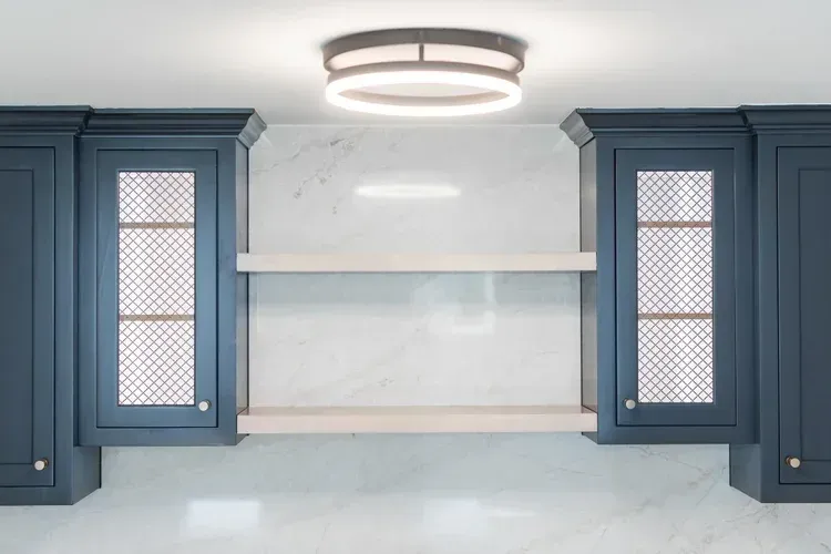 Blue kitchen cabinets framing a white marble backsplash and floating shelves under a modern ceiling light