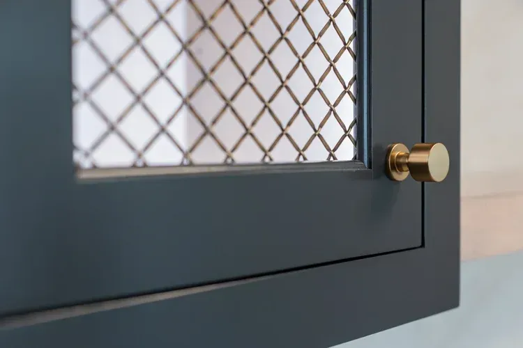 Close-up of a dark gray cabinet door with wire mesh panel and brass knob