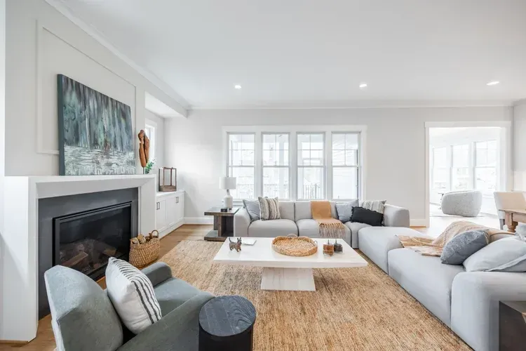Bright living room with gray sofas, white fireplace, large windows, and a woven rug