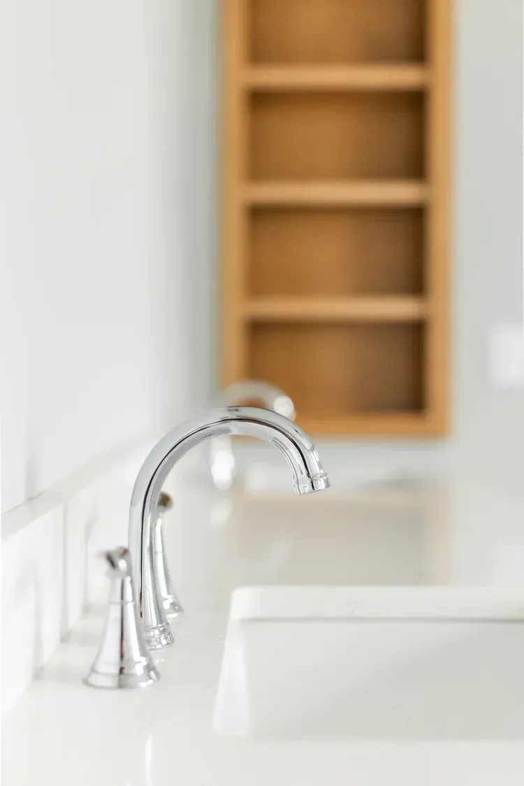 Chrome faucet over a white sink with a blurred wooden shelf in the background