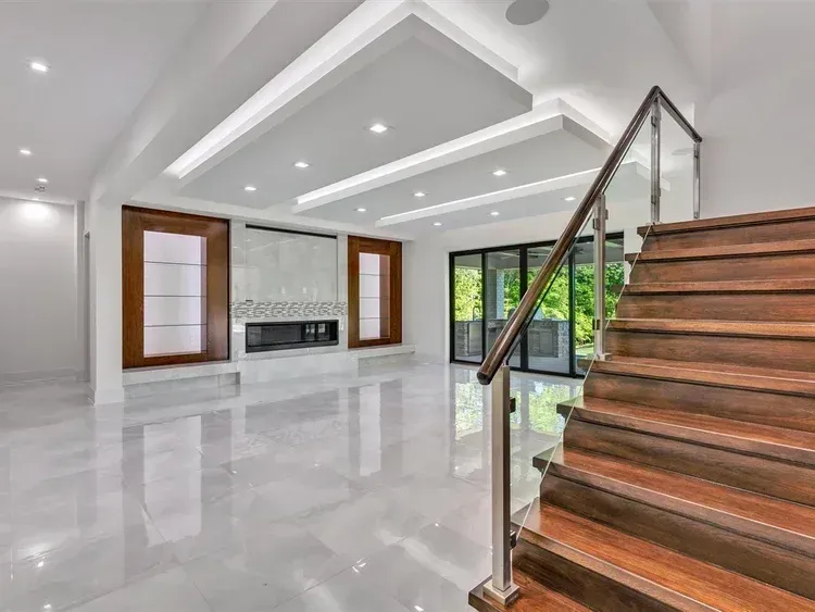 Modern open living room with glossy tile floors, recessed lighting, and a wooden staircase with glass railing