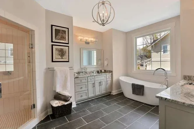 Bright bathroom with freestanding tub, large window, gray tile floor, and vanity with mirror.
