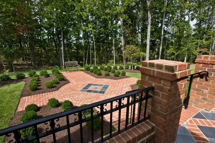 Brick patio and garden path bordered by shrubs, with trees and a black metal railing in the foreground.