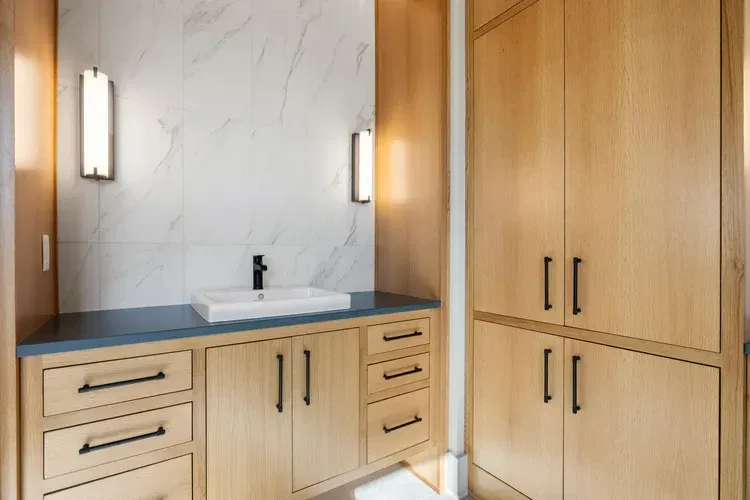 Modern bathroom vanity with light wood cabinets, blue countertop, marble wall, and two wall sconces