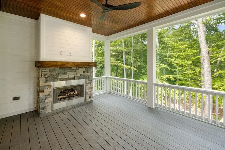 Covered porch with stone fireplace, gray floor, white railings, and wooded view beyond large windows